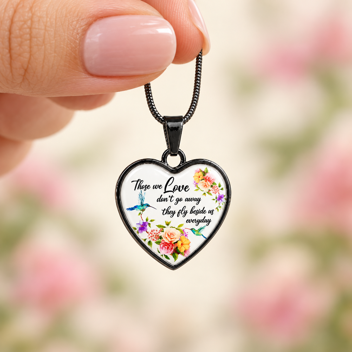 Woman’s hand holding a small black heart-shaped hummingbird memorial necklace against a softly blurred floral background.