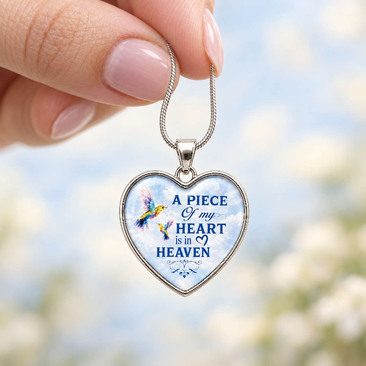Close-up of a woman’s hand holding a small heart-shaped hummingbird memorial necklace against a softly blurred blue background.