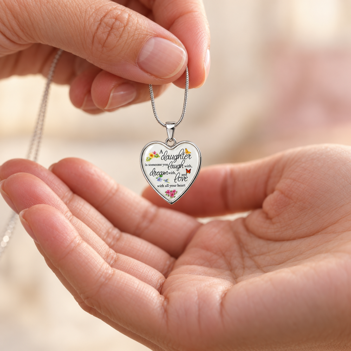 A close-up lifestyle shot of a person's hands holding a silver heart-shaped pendant necklace. The pendant features the words 'Daughter laugh prosper love' in black script, decorated with small colorful floral illustrations, and is suspended from a delicate silver chain against a soft, blurred background.