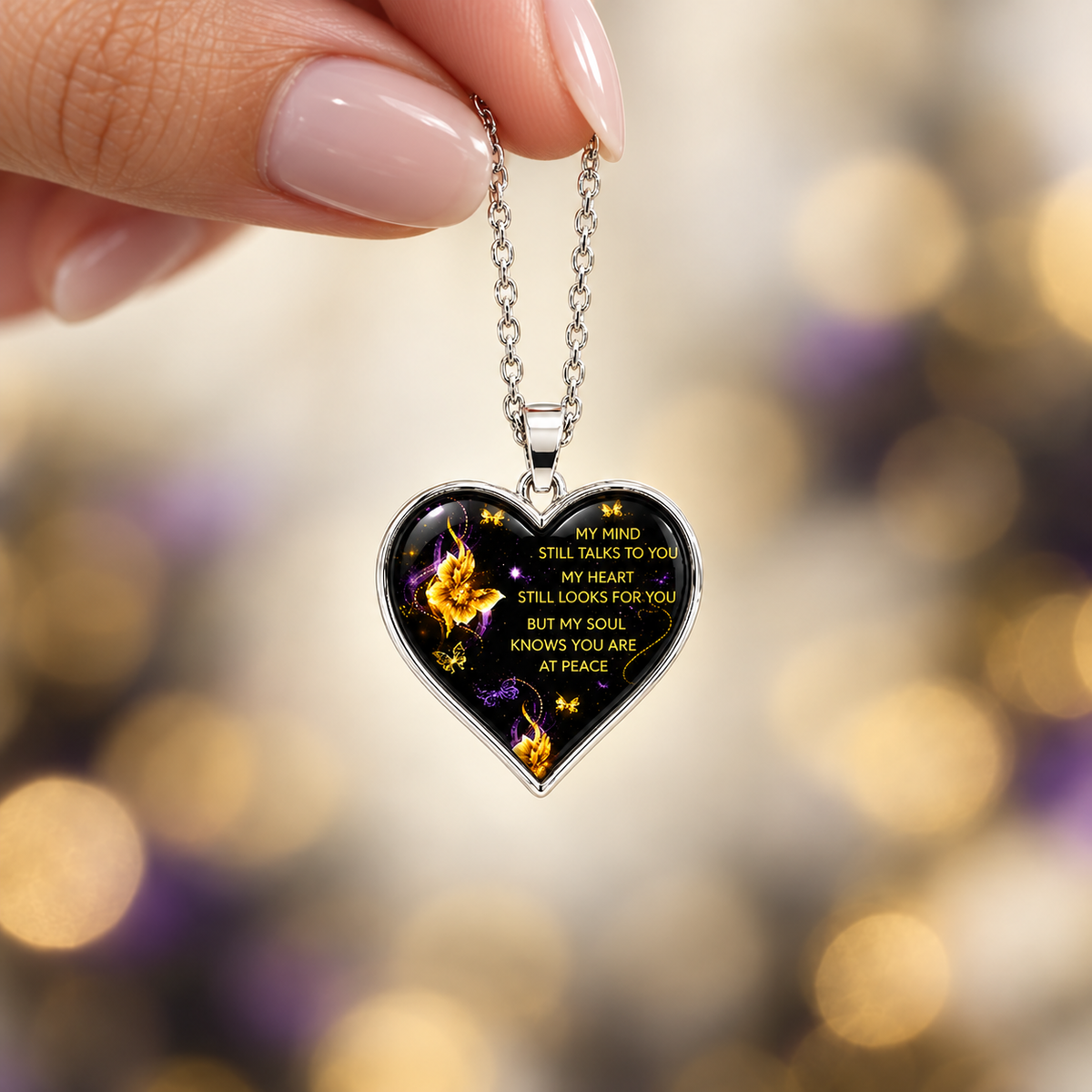 Woman’s hand holding a small black and silver memorial heart necklace on a linked chain against a softly blurred ivory, purple and gold background.