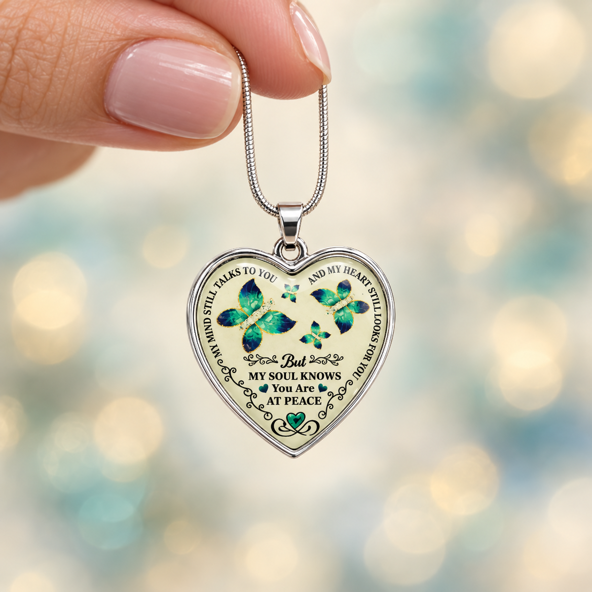Woman’s hand holding a small silver butterfly memorial necklace against a softly blurred ivory, teal and champagne background.