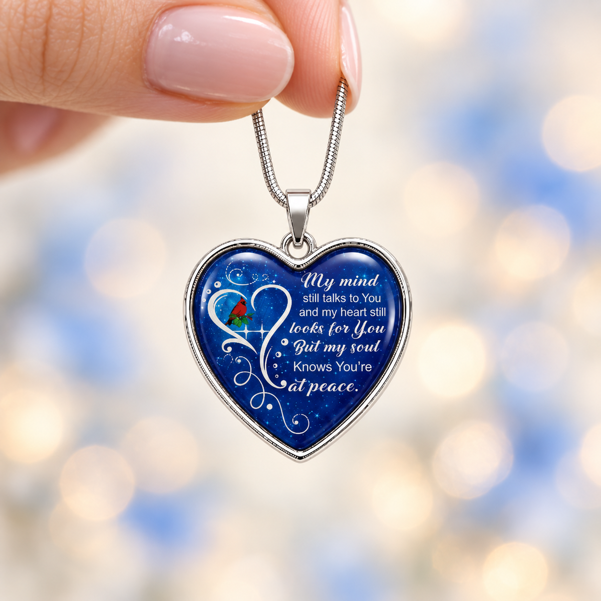 Woman’s hand holding a small silver cardinal memorial necklace against a softly blurred ivory, pale-blue and champagne background.