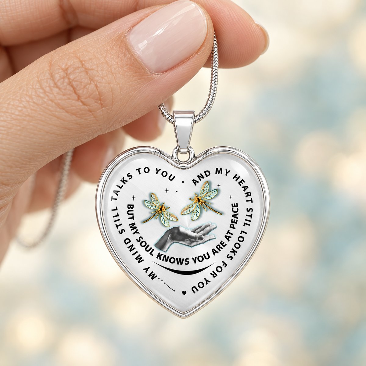 A close-up of a hand holding a silver heart-shaped pendant necklace. The pendant has a white background with black text, a small illustration of a hand, and two green dragonflies, set against a soft, blurred bokeh background.