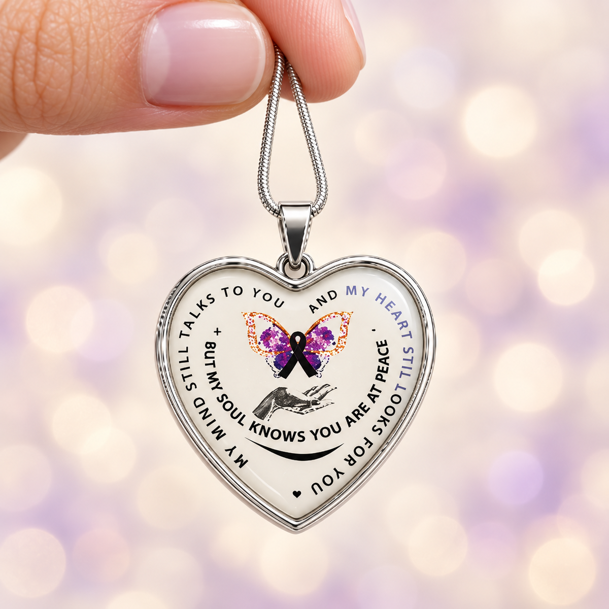 A silver heart-shaped pendant necklace held by a hand, featuring a white background with black text, a purple and orange butterfly, and a black awareness ribbon, set against a soft purple bokeh background.