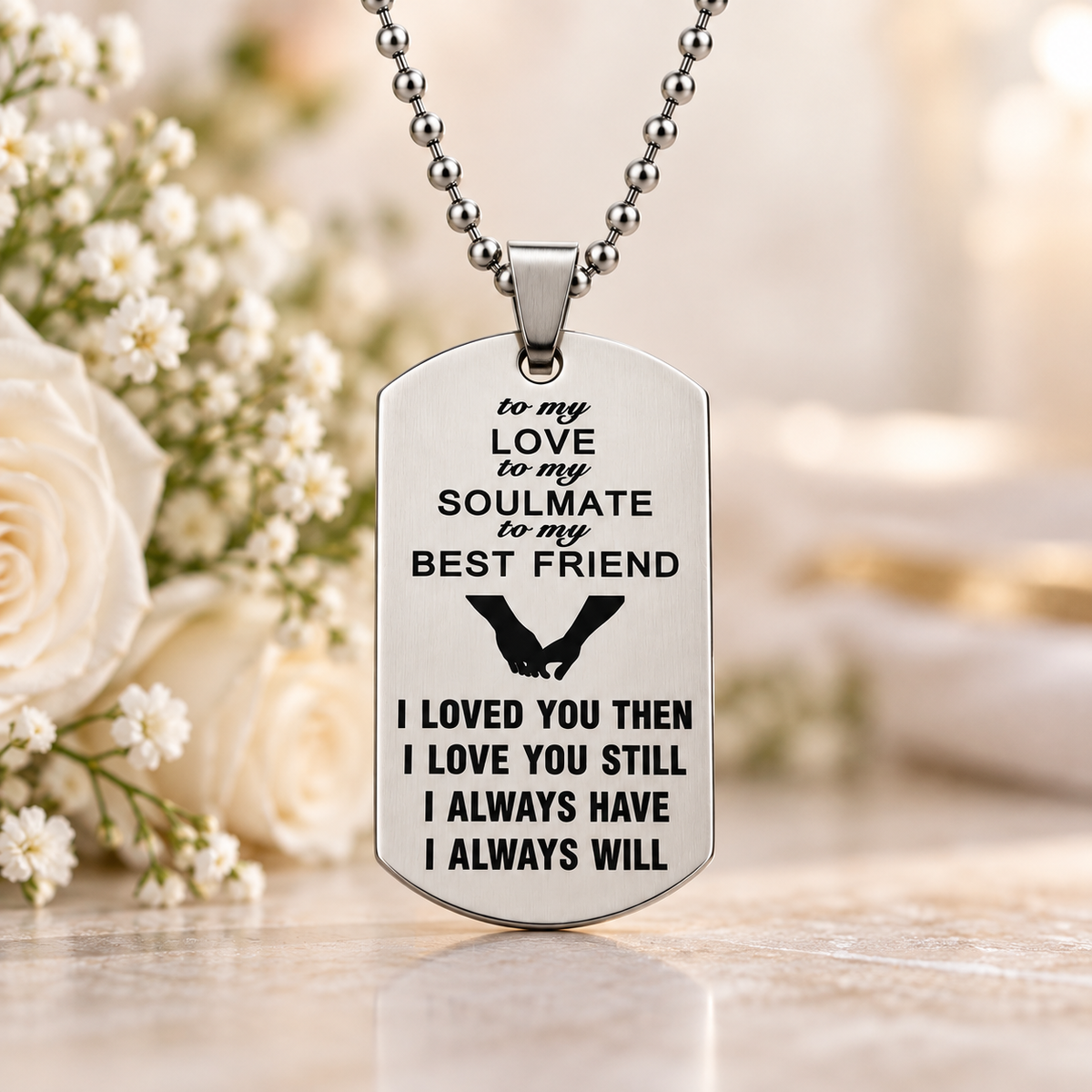 A silver-toned dog tag necklace with engraved black text and a heart design, displayed on a marble surface with white roses in the background.