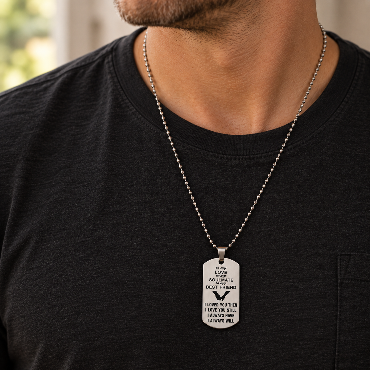 A close-up of a man wearing a silver ball-chain necklace with a dog tag pendant engraved with a message for a best friend, styled with a black t-shirt.
