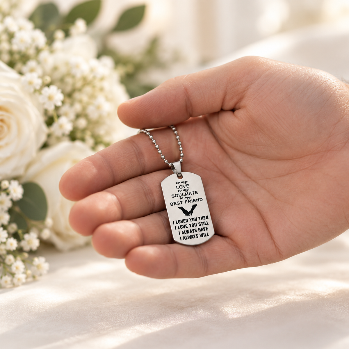 A hand holding a silver ball chain necklace with a white dog tag pendant engraved with 'I LOVE YOU SOULMATE BEST FRIEND' and a heart design, with white flowers blurred in the background.