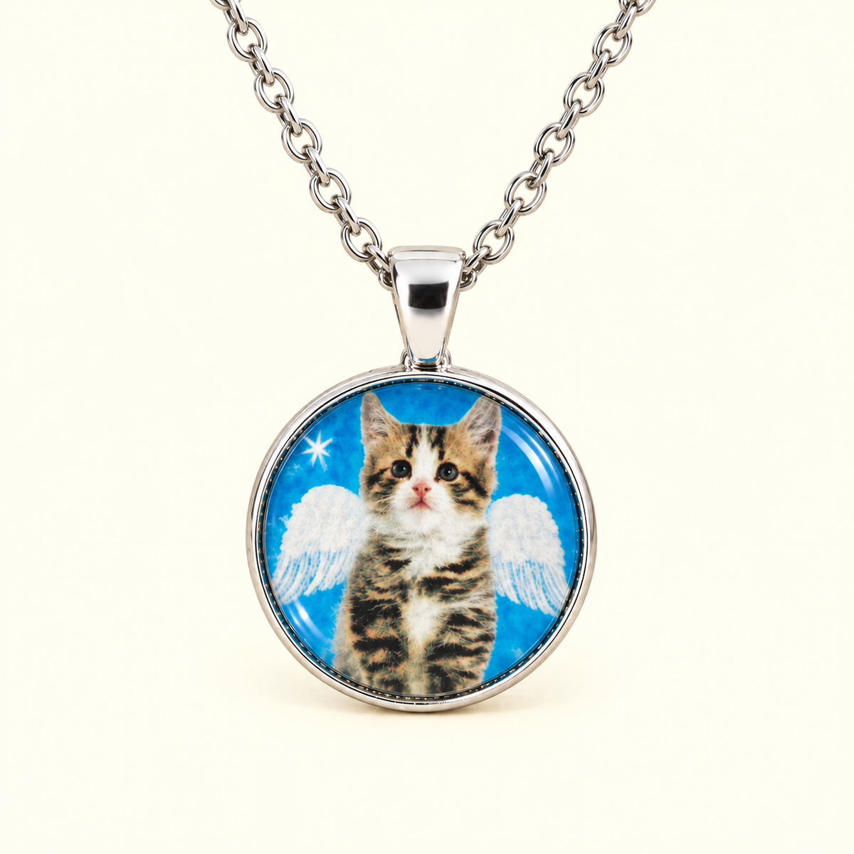 A silver chain necklace with a round pendant featuring a printed photo of a kitten with white angel wings against a bright blue background, shown on a plain light cream background.