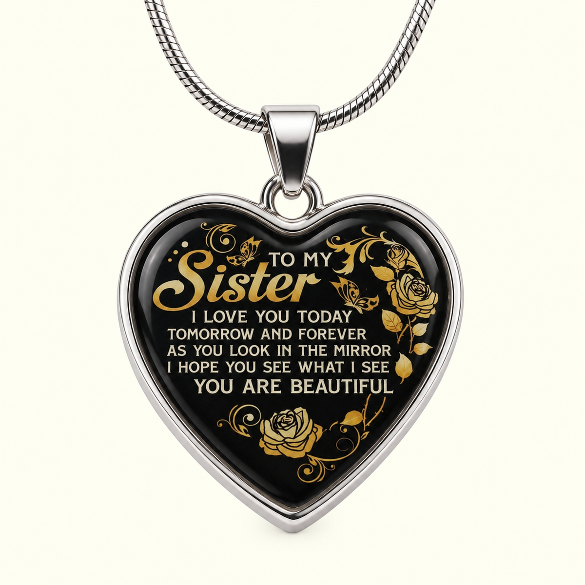 A heart-shaped pendant necklace with a silver-tone border and chain, featuring a black face with gold floral designs and a sentimental quote for a sister, shown on a light cream background.