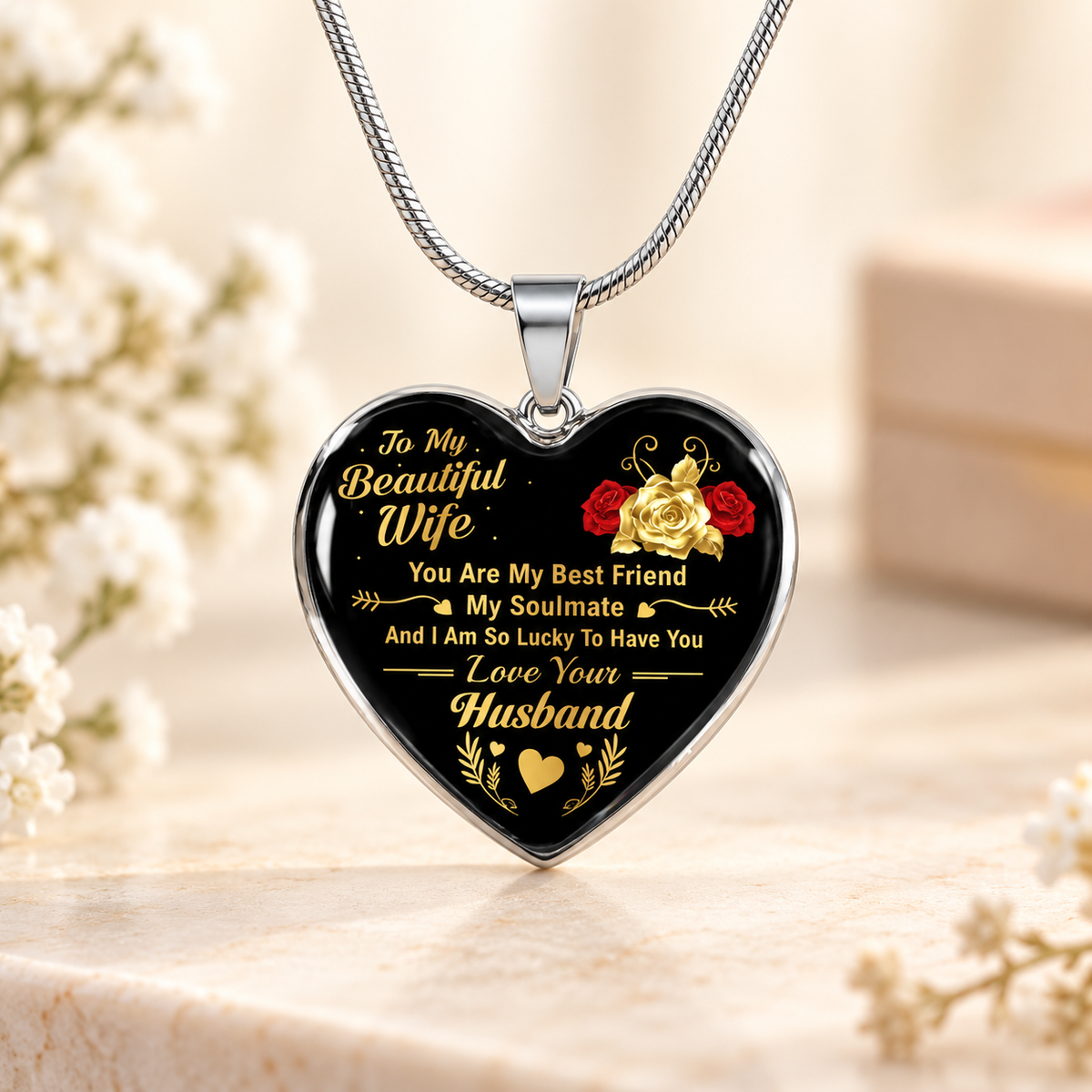 A heart-shaped pendant necklace with a black background and silver border, featuring gold and red text and rose graphics, hanging on a silver chain against a softly blurred beige background with white flowers.