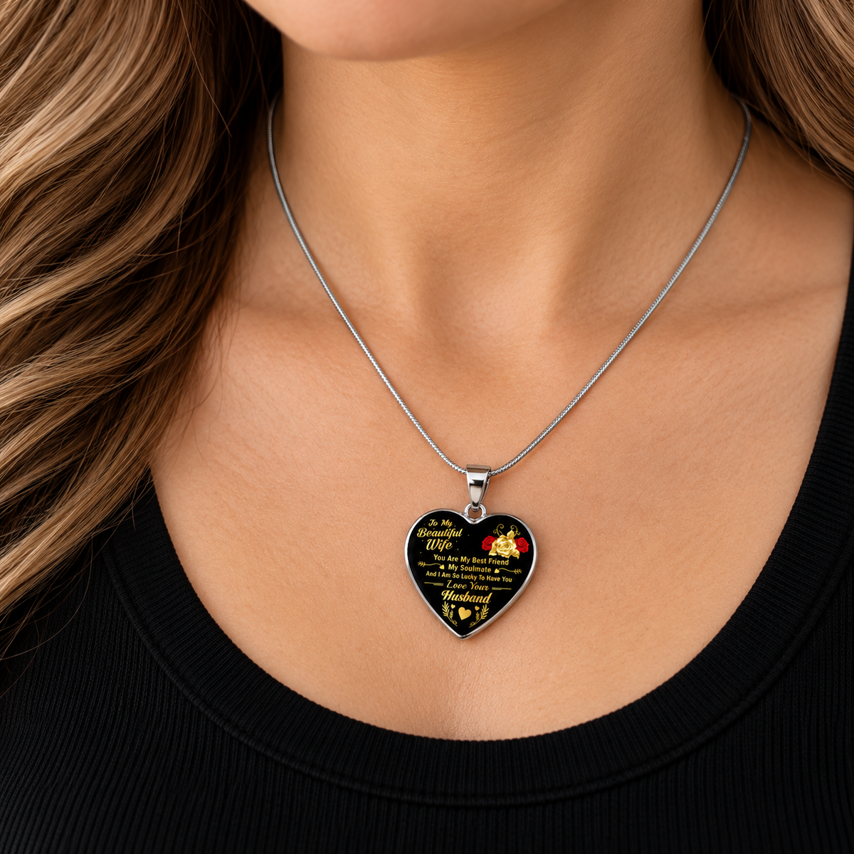 A close-up of a woman wearing a heart-shaped pendant necklace. The pendant is black with gold text and a small red floral accent, suspended from a silver chain.