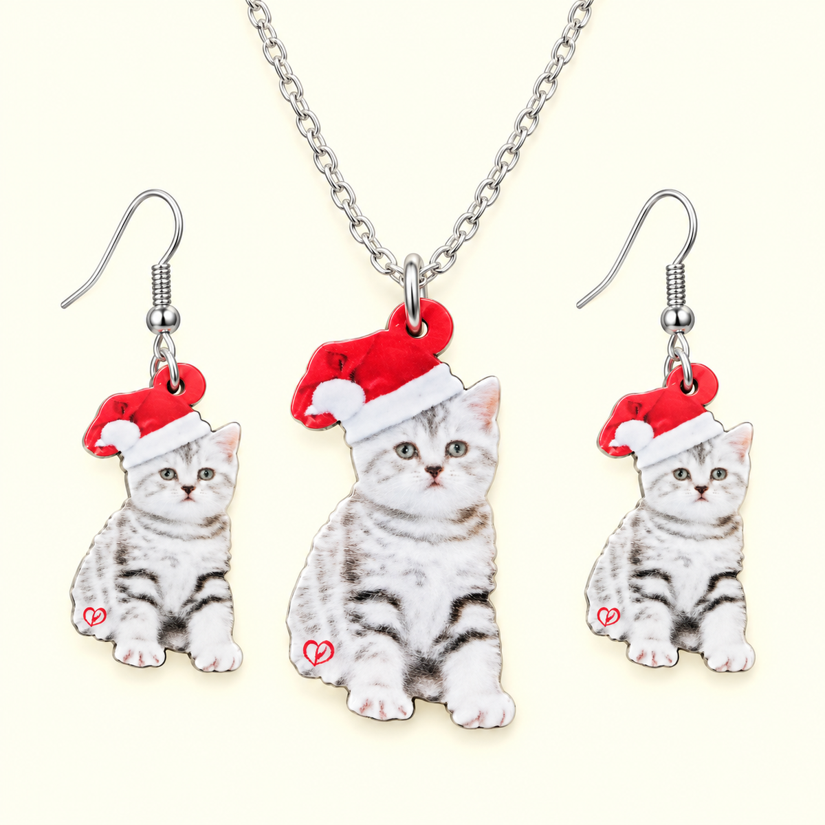 A matching jewelry set featuring a silver chain necklace and two dangling earrings, each with a charm shaped like a gray tabby kitten wearing a red Santa hat. The kitten charms have printed fur details and a small red heart on the side, set against a light cream background.