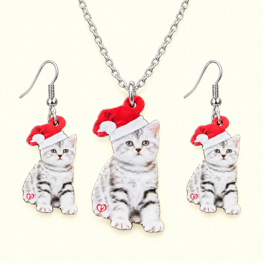 A matching jewelry set featuring a silver chain necklace and two dangling earrings, each with a charm shaped like a gray tabby kitten wearing a red Santa hat. The kitten charms have printed fur details and a small red heart on the side, set against a light cream background.