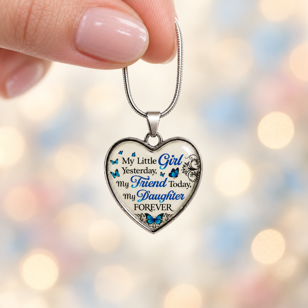 Woman’s hand holding a small silver daughter heart necklace with blue butterflies against a softly blurred ivory, blush and pale-blue background.
