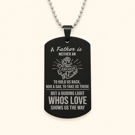 A black dog tag pendant necklace with a silver ball chain, featuring engraved white text and an anchor graphic on a light cream background.