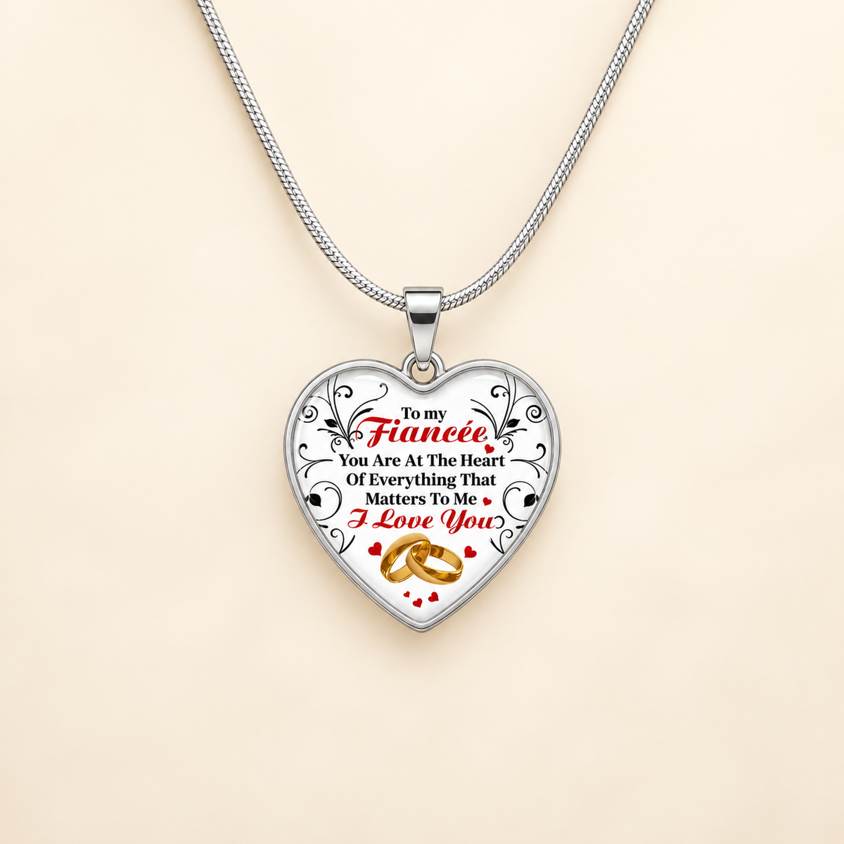 A silver heart-shaped pendant necklace on a matching silver chain, featuring a printed message for a fiancée with red and black text and gold wedding rings, displayed on a light beige background.
