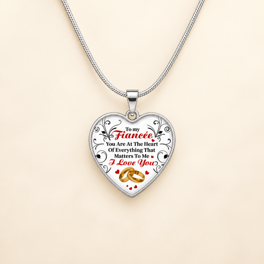 A silver heart-shaped pendant necklace on a matching silver chain, featuring a printed message for a fiancée with red and black text and gold wedding rings, displayed on a light beige background.