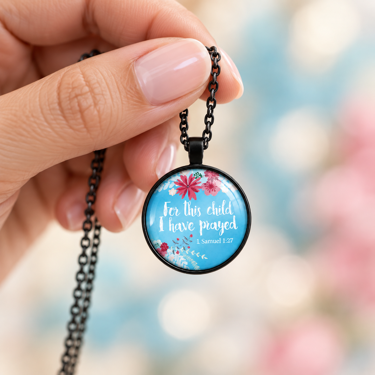 Woman’s hand holding a round black necklace with blue floral artwork and the words “For this child I have prayed” from 1 Samuel 1:27.