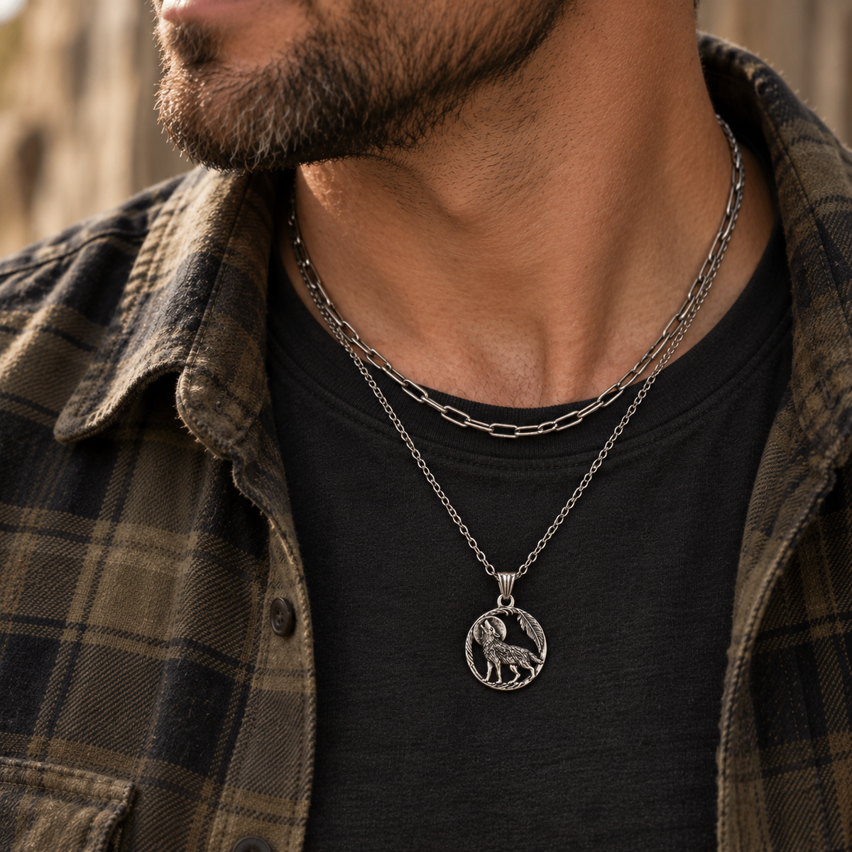 A close-up of a man wearing a silver pendant necklace with a detailed animal emblem, layered over a black t-shirt and a brown plaid flannel shirt.