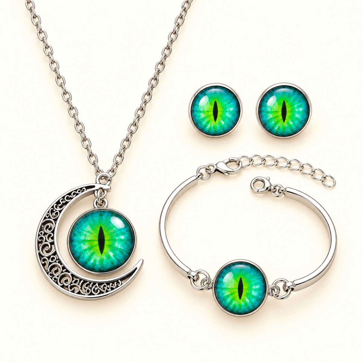 A three-piece gothic jewelry set featuring a silver crescent moon necklace, a matching silver bangle bracelet, and a pair of stud earrings, all adorned with vibrant green and blue reptile eye cabochons on a light cream background.