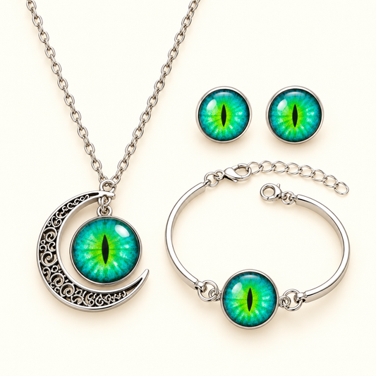 A three-piece gothic jewelry set featuring a silver crescent moon necklace, a matching silver bangle bracelet, and a pair of stud earrings, all adorned with vibrant green and blue reptile eye cabochons on a light cream background.