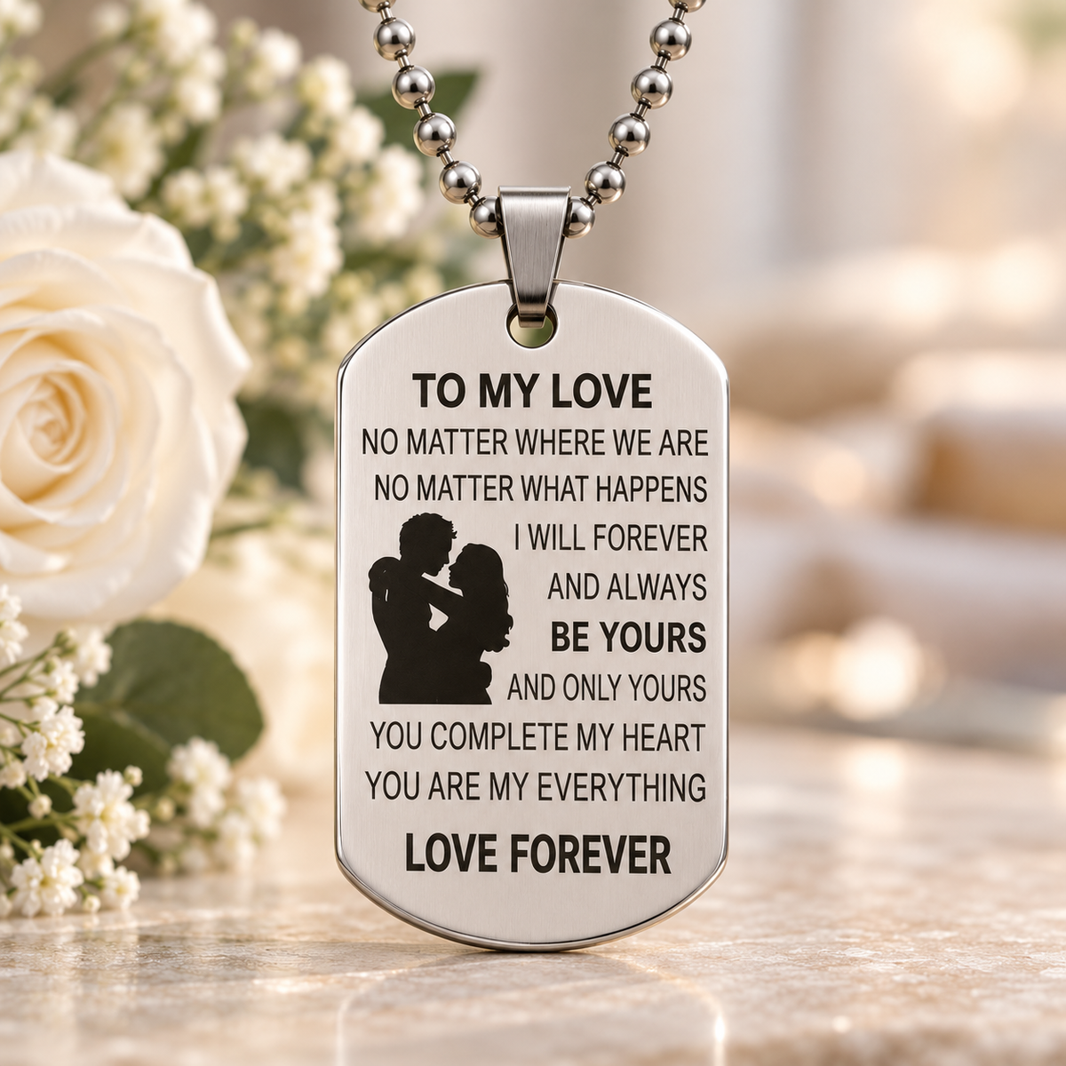 A silver-toned dog tag necklace with engraved sentimental text and a silhouette of a couple, hanging on a ball chain. The pendant is displayed on a reflective surface with a blurred background featuring a white rose and soft warm lighting.