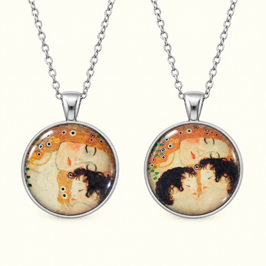 Two matching silver chain necklaces with round pendant frames featuring a vintage-style illustration of two embracing figures in warm beige, orange, and black tones, shown on a light cream background.