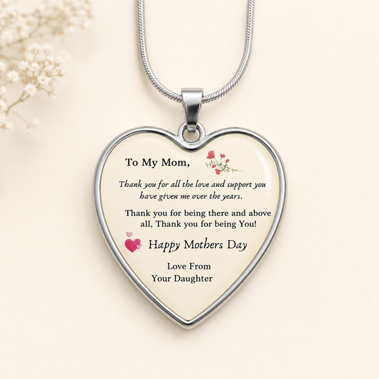 A heart-shaped pendant necklace with a silver-toned metal border and chain, featuring a printed Mother's Day thank-you message on a cream background with small pink heart and floral accents, shown on a light beige backdrop.