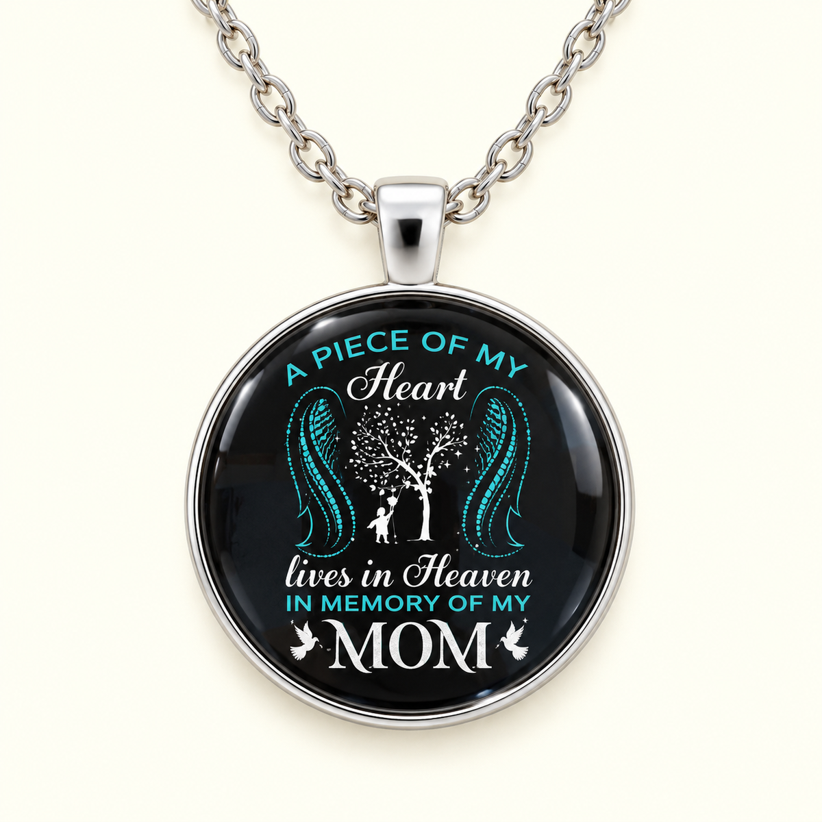 A silver round pendant necklace with a black background, featuring a printed design of a tree with teal wings and the text 'A PIECE OF MY Heart lives in Heaven IN MEMORY OF MY MOM'.