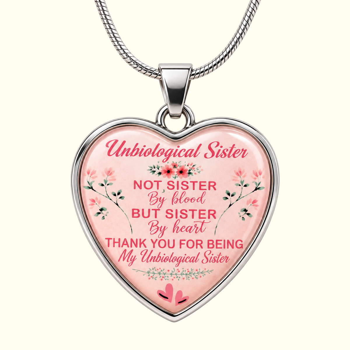 A heart-shaped pendant necklace with a silver-toned metal frame and chain, featuring a pink background with floral illustrations and the text 'Unbiological Sister NOT SISTER By blood BUT SISTER By heart THANK YOU FOR BEING My Unbiological Sister' on a light cream background.
