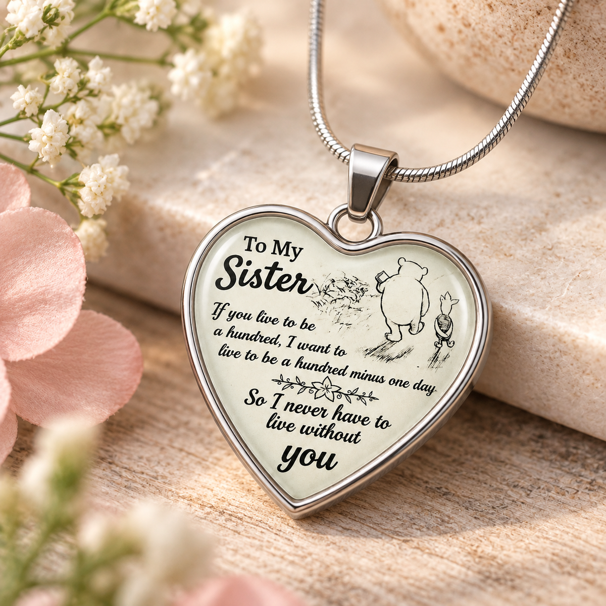 image of a heart shaped pendant with pooh and piglet which reads to my sister if you live to be a hundred, i want to live to be a hundred minus one day so I never have to live without you sitting on a wooden table with pink and white flowers