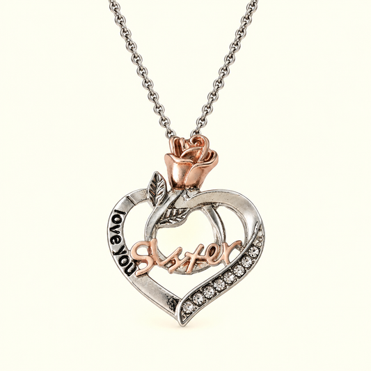 A silver and rose gold heart pendant necklace with a rose at the top, small clear rhinestones along one edge, and the words 'I love you sister' engraved on the pendant, shown on a light cream background.