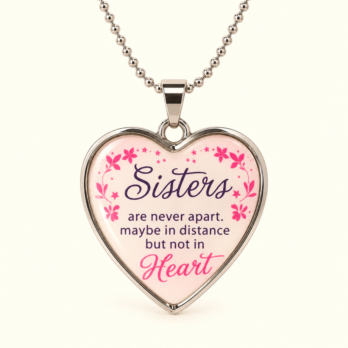 A heart-shaped pendant necklace with a silver-toned metal border and ball chain, featuring a light pink background with the text 'Sisters are never apart. maybe in distance but not in Heart' in purple and pink script, decorated with small pink flowers and stars.